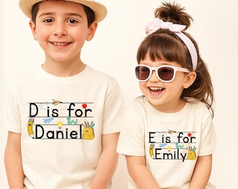 May include: Two off-white short-sleeved t-shirts featuring school-themed graphics. One shirt displays the text 'D is for Daniel' with illustrations of a pencil, apple, paper airplane, and backpack. The other shirt displays 'E is for Emily' with identical icons.