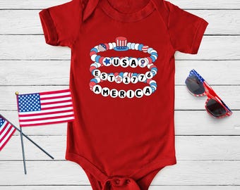 Puede incluir: Un body de bebé rojo de manga corta con un gráfico de tres pulseras de cuentas apiladas en rojo, blanco y azul. Las cuentas forman las palabras USA, EST 1776 y AMERICA, decoradas con un pequeño icono de sombrero de copa y formas de estrellas.