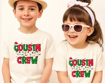 May include: Two off-white short-sleeve t-shirts featuring the text COUSIN CREW in red and green letters. The design includes a string of colorful Christmas lights, small snowflakes, and a red Santa hat perched on the letter C.