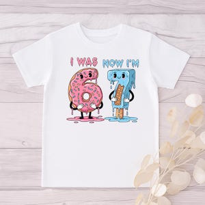 May include: White t-shirt with a colorful graphic. The graphic features the text "I WAS NOW I'M" above the numbers 6 and 7, designed as a donut and an ice cream bar. The donut is pink, and the ice cream bar is blue.