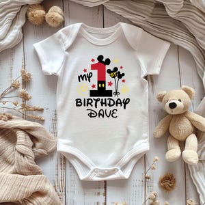 K&ouml;nnte beinhalten: Wei&szlig;er Baby-Body mit farbenfroher Grafik zum ersten Geburtstag. Das Design umfasst eine gro&szlig;e rote Zahl 1, Mickey Mouse Ohren, Sterne und den Text "my 1st birthday Dave". Ein Teddyb&auml;r liegt neben dem Body.