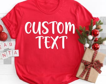 Puede incluir: Una camiseta roja con las palabras "CUSTOM TEXT" en escritura blanca. La camiseta se muestra con decoraciones navideñas, incluyendo adornos, un regalo envuelto y vegetación. La camiseta es un artículo festivo.