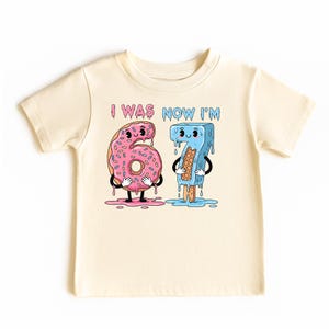 May include: Cream-colored toddler t-shirt with a graphic featuring the numbers 6 and 7 as cartoon characters. The number 6 is a pink donut, and the number 7 is a blue popsicle. The text "I WAS NOW I'M" is above the numbers.