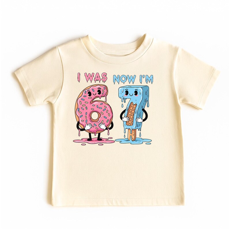 May include: Cream-colored toddler t-shirt with a graphic featuring the numbers 6 and 7 as cartoon characters. The number 6 is a pink donut, and the number 7 is a blue popsicle. The text "I WAS NOW I'M" is above the numbers.