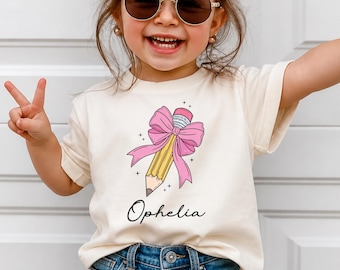 May include: A cream-colored short-sleeve t-shirt featuring a graphic of a yellow pencil adorned with a large pink bow. The name Ophelia is printed in black cursive script below the pencil graphic. The design includes small black star accents.
