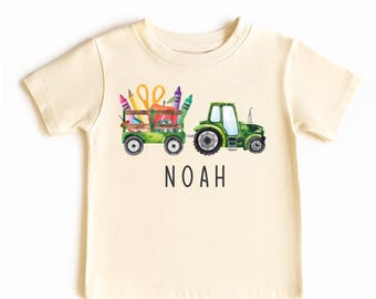 May include: A cream-colored short-sleeve t-shirt featuring a watercolor graphic of a green tractor pulling a wooden wagon filled with school supplies including crayons, a pencil, scissors, and an apple, with the name NOAH printed below.