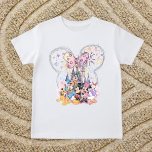 A white t-shirt with a colorful graphic design featuring Disney characters Mickey Mouse, Donald Duck, and Goofy, along with a castle and other whimsical elements.