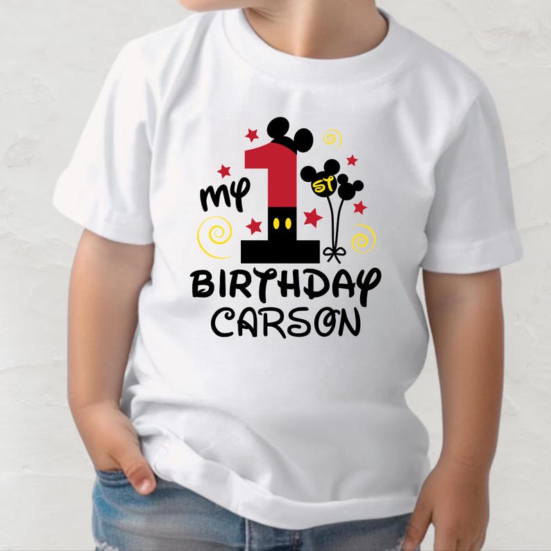K&ouml;nnte beinhalten: Wei&szlig;es T-Shirt mit einem farbenfrohen Grafikdesign f&uuml;r einen ersten Geburtstag. Das Design umfasst eine gro&szlig;e rote Zahl 1, Mickey Mouse Ohren, Sterne und den Text "my 1st Birthday Carson."