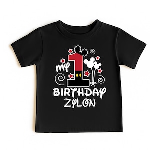 K&ouml;nnte beinhalten: Schwarzes Kleinkind-T-Shirt mit einer farbenfrohen Grafik zum ersten Geburtstag. Das Design umfasst eine gro&szlig;e rote Zahl 1, Mickey Mouse Ohren, Sterne und den Text "My Birthday Zylon."