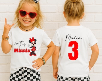 May include: A white t-shirt featuring a graphic of Minnie Mouse with the text i'm this Minnie on the front and the name Malia with the number 3 in red on the back. The shirt is paired with black and white checkered shorts and a matching hair bow.