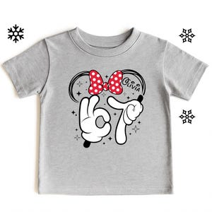 A gray t-shirt with a graphic design featuring a red bow, a peace sign, and the text "Olivia" along with a snowflake.