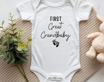 May include: White baby onesie with the text "FIRST Great Grandbaby" in black script. Below the text are two black footprints. The onesie is on a beige textured surface, with a stuffed animal and greenery in the background.