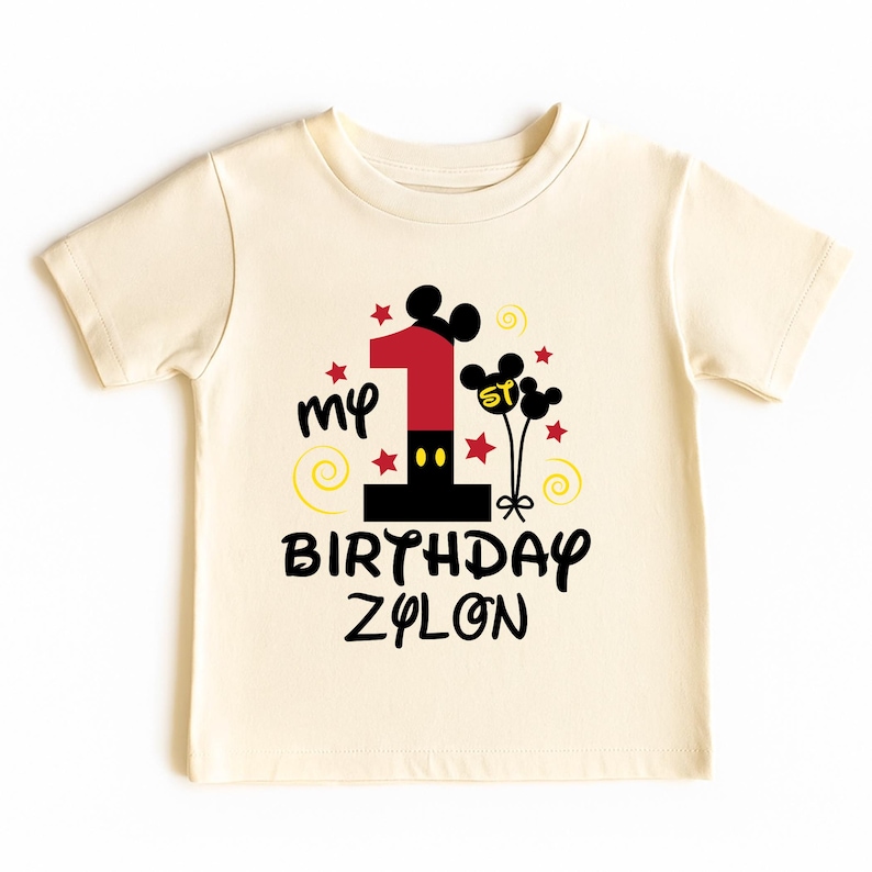 K&ouml;nnte beinhalten: Cremefarbenes Kleinkind-T-Shirt mit einer farbenfrohen Grafik. Das Design zeigt eine gro&szlig;e rote Zahl 1, Mickey Mouse Ohren, Sterne und die Worte "my BIRTHDAY ZYLON" in Schwarz. Gelbe Wirbel und Mickey Mouse Ballons vervollst&auml;ndigen den festlichen Look.