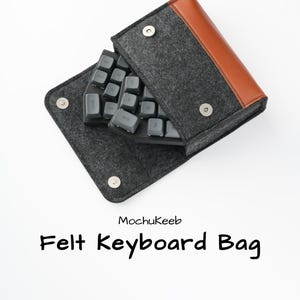 Könnte beinhalten: Eine graue Filz-Tastaturtasche mit braunem Lederakzent und Druckknöpfen. Die Tasche enthält eine graue Tastatur mit schwarzen Tasten. Der Text "MochuKeeb" und "Felt Keyboard Bag" sind darunter gedruckt.