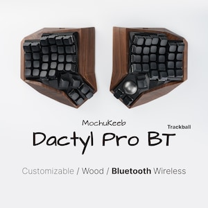 May include: A split ergonomic keyboard with dark wood housing and black keys. The right half features a silver trackball. Text reads: Mochukeeb Dactyl Pro BT Trackball Customizable / Wood / Bluetooth Wireless. The design is angled and contoured.