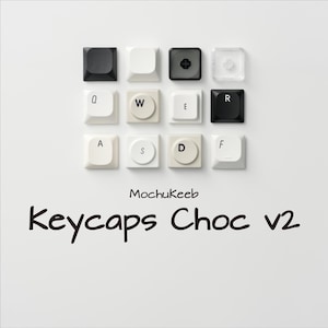 Op de afbeelding: Een bovenaanzicht van een verzameling vierkante keycaps in verschillende kleuren, waaronder zwart, wit en transparant. Sommige keycaps hebben letters erop gedrukt. De tekst "MochuKeeb Keycaps Choc v2" staat eronder.