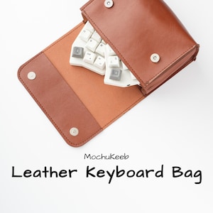 May include: A brown leather keyboard bag with a white keyboard partially visible inside. The bag is open, revealing a tan interior and silver snap closures. The text "MochuKeeb" and "Leather Keyboard Bag" are printed below.