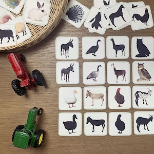Può includere: Flashcard di animali con illustrazioni di animali da fattoria, come asini, oche e mucche, disposte su una superficie di legno. Le carte sono quadrate e bianche con silhouette nere. Sono presenti anche trattori giocattolo e un cestino.