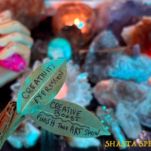 Puede incluir: Primer plano de una varita de hojas con las palabras "Creativity Expression" y "Creative Boost Finish That Art Show" escritas en las hojas. El fondo incluye cristales, velas y el texto "Shasta Spells".