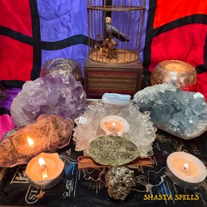 Puede incluir: Una colección de cristales, velas y una jaula para pájaros en una bandeja. Los cristales incluyen amatista y celestita. Un letrero dice "Creative Boost for Songwriters". La bandeja tiene el texto "Shasta Spells."