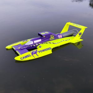 May include: A radio-controlled hydroplane boat in bright neon yellow and purple. The hull features black and white checkered patterns, the text "CAMEL POWERED" with a camel silhouette, "Joe's Racing" branding, and a rear spoiler on the water.