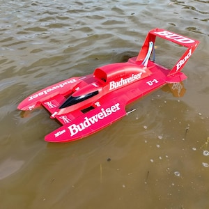May include: A red remote-controlled hydroplane boat floating on water. The boat features the Budweiser logo in white text on the sides and rear wing, with the text Miss Budweiser and U-1 visible. It has a black cockpit canopy and a sleek design.