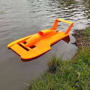 May include: A bright orange remote-controlled hydroplane boat floating on water. The model features a flat, wide hull design with a central cockpit cowl, a rear-mounted engine housing, and a tall, angular rear stabilizer wing structure.