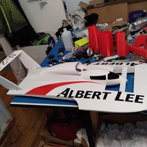 May include: A white remote-controlled hydroplane boat model featuring red accents and black text that reads ALBERT LEE on the side and U-21 on the rear stabiliser fin. The model has a sleek aerodynamic hull with a black cockpit canopy window.