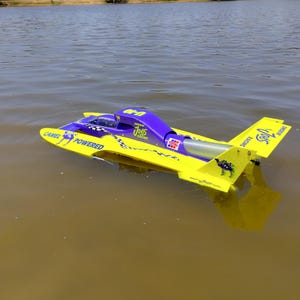 May include: A bright yellow and purple remote-controlled hydroplane racing boat floating on water. The boat features the text Camel Powered, U-10, and Smokin Joes Racing, along with a checkered flag pattern and a small camel graphic on the side.