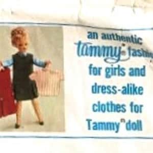 Può includere: Una bambola con un vestito blu e bianco tiene in mano due vestiti aggiuntivi, uno rosso e uno rosa e bianco a righe. Il testo sulla confezione recita "una autentica moda Tammy per ragazze e abiti uguali per la bambola Tammy."