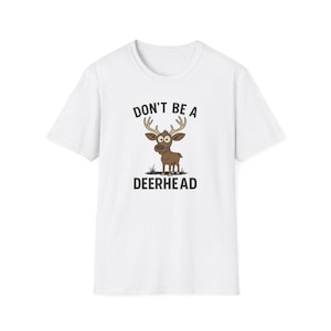 Puede incluir: Camiseta blanca con un ciervo de dibujos animados y el texto "DON'T BE A DEERHEAD". El ciervo es marrón con ojos grandes y cuernos. Una prenda de vestir casual.