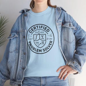 May include: Pale blue t-shirt featuring a black and white circular graphic with the text "CERTIFIED PROBLEM SOLVER" and mathematical symbols. The shirt is made of a soft material. The t-shirt is a casual wear item.