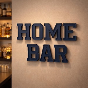 May include: Dark blue "HOME BAR" sign in a bold, block letter font, mounted on a tan wall. The letters are three-dimensional, creating a shadow effect. A bar with bottles is visible in the background.