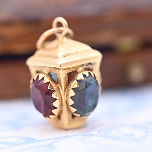 Könnte beinhalten: Ein goldfarbener Laternenanhänger mit roten und blauen Edelsteinen. Der Anhänger hat ein detailliertes Design mit einer Schlaufe oben für eine Kette. Die Edelsteine sind facettiert und in einem dekorativen Goldrahmen gefasst.
