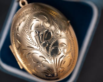 Könnte beinhalten: Ein goldfarbenes ovales Medaillon mit aufwendigen floralen und Blattgravuren. Das Medaillon ist geöffnet und gibt einen Blick ins Innere frei. Es liegt in einer blau samtgefütterten Schachtel, was auf eine Präsentation oder Aufbewahrung hindeutet.