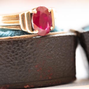 Könnte beinhalten: Ein goldener Ring mit einem großen, oval geschliffenen Rubin. Der Ring ist in einem klassischen Design gefasst und liegt in einer Vintage-Schmuckschatulle in Dunkelbraun mit türkisfarbenem Innenraum. Die Schatulle ist geöffnet und zeigt den Ring.