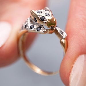 May include: A gold ring with a leopard head design, embellished with black and clear gemstones. The ring is held up, highlighting the detailed craftsmanship. The leopard's head is the focal point, with a gold-toned band. The ring's design is intricate and eye-catching.