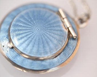 May include: Close-up of a light blue and silver pendant necklace. The pendant is circular with a detailed radial pattern and a hinged lid. The necklace is a delicate piece of jewelry.