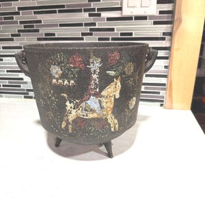May include: A dark, cast iron cauldron with three legs and two handles. The cauldron is decorated with a painted scene featuring a horse and rider, with red and white floral accents. The cauldron is likely used for decorative purposes.