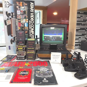 May include: An Atari Video Computer System console with a wood-grain front, a small television displaying gameplay, two black joysticks, a power adapter, multiple game cartridges, an original box, an owner's manual, and a product catalog.