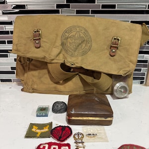 May include: A tan canvas Boy Scouts of America messenger bag with two brass buckles, accompanied by a collection of scouting memorabilia including a wooden box, a compass, embroidered patches with text like 209 and Be Prepared, and pins.