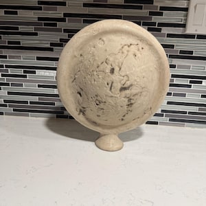 May include: A beige, round decorative object with a textured surface and a pedestal base. The object has a raised, circular design on its face. The background features a white countertop and a mosaic tile backsplash.