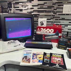 May include: An Atari 2600 video game console with a black and silver design, a red box, and several game cartridges. The console is next to a black television displaying a game screen. Game manuals are also visible.
