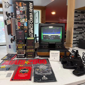May include: An Atari 2600 video computer system console with a wood-grain front, a television screen displaying gameplay, two black joysticks, a stack of game cartridges, the original box, an owner's manual, a catalog, and a power adapter box.