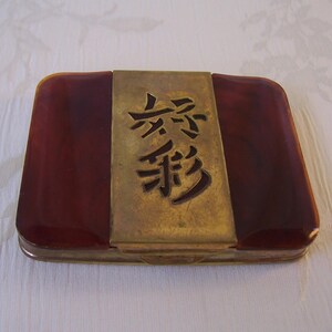 May include: A rectangular compact case featuring a central brass panel with dark cut-out characters. The sides are made of a smooth, deep reddish-brown material with a marbled appearance. The metal has a muted, aged finish with visible patina.