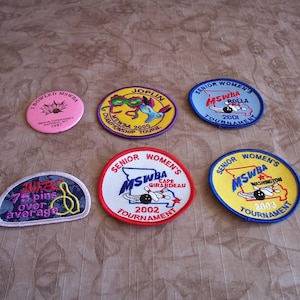 May include: A collection of bowling patches and pins. The patches feature text such as "MSWBA", "Senior Women's Tournament", and various city names. Colors include pink, blue, yellow, and red. The pin reads "I Bowled MSWBA 1997".