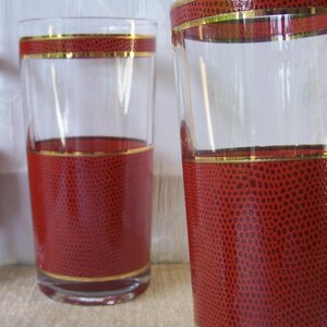 May include: Two clear glass tumblers featuring a wide band of textured red material with a dotted pattern around the lower section and a thinner matching band near the rim, both accented by thin horizontal gold-colored metallic stripes.