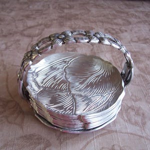 May include: A silver-colored serving basket with a floral handle and a stack of matching plates. The plates have a textured, leaf-like design. The handle is adorned with detailed flower accents.