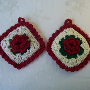 May include: Two square crochet potholders featuring a central three-dimensional red rose motif surrounded by green leaves. The base is off-white with a scalloped red border and a small loop at the top for hanging. The design is identical on both items.