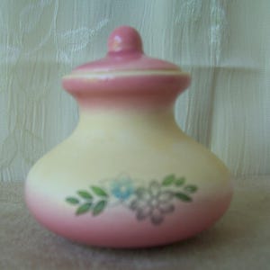 May include: A small ceramic jar with a rounded body and a matching lid. The jar features a gradient color scheme, transitioning from a soft pink at the base and lid to a pale yellow in the center. A floral design with green leaves is painted on the front.